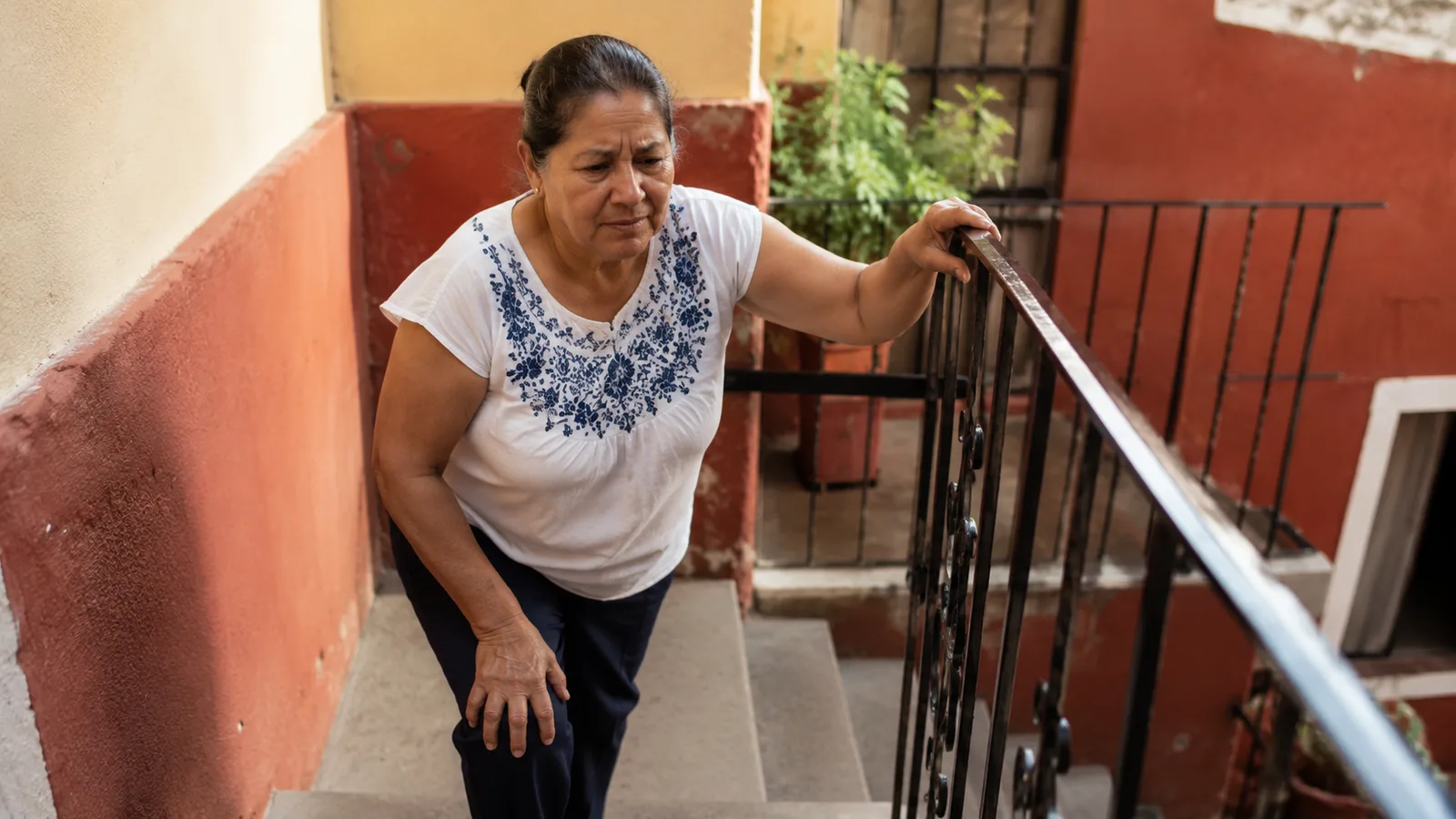 Mujer mexicana mayor subiendo escaleras con apoyo en el barandal por dolor de rodilla